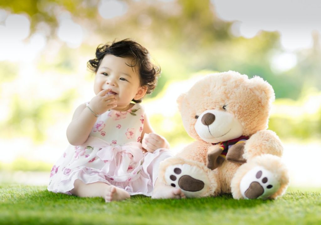 Baby sitting on grass beside bear plush toy at daytime