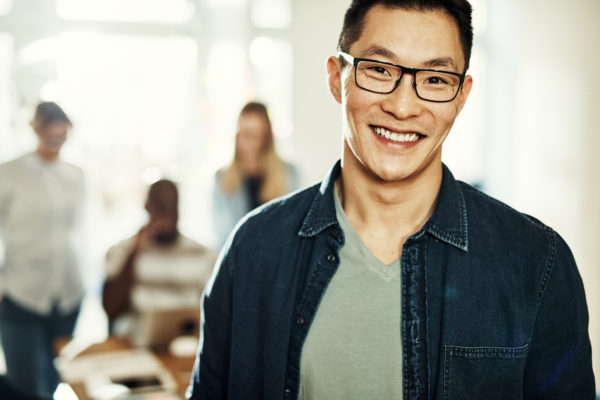 Young Asian designer wearing glasses and smiling confidently while standing in a modern office with colleagues in the background